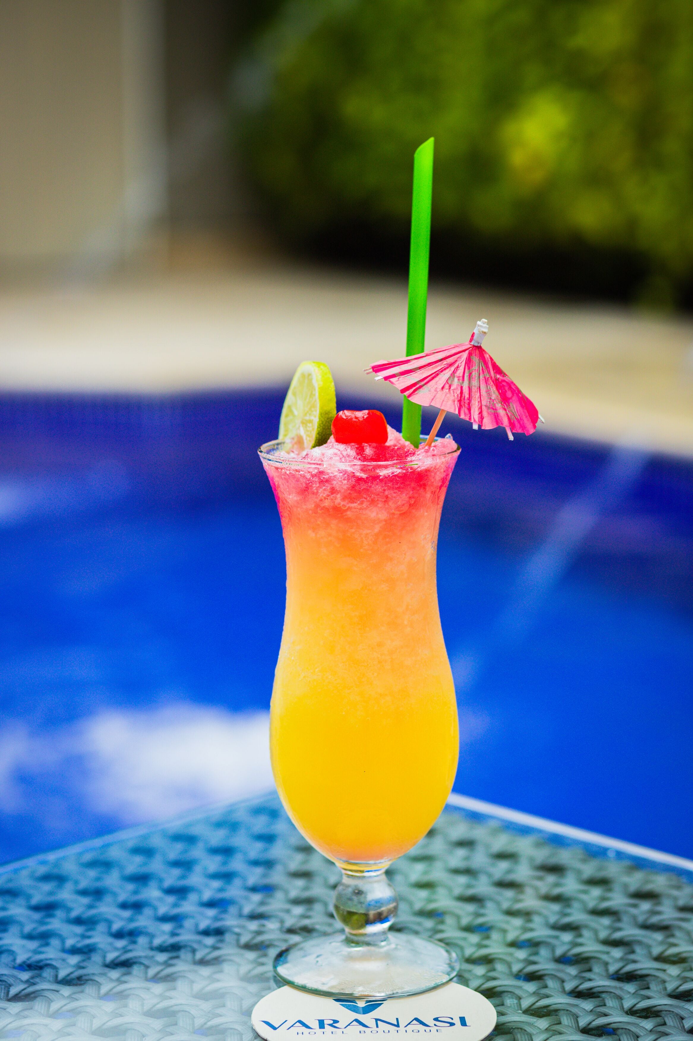 Um cocktail num copo alto, com uma fatia de limão e um guarda-sol cor-de-rosa, sobre uma mesa junto a uma piscina.