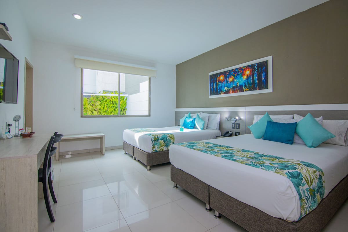 A hotel room with two beds, a desk, and a window with a view of greenery.