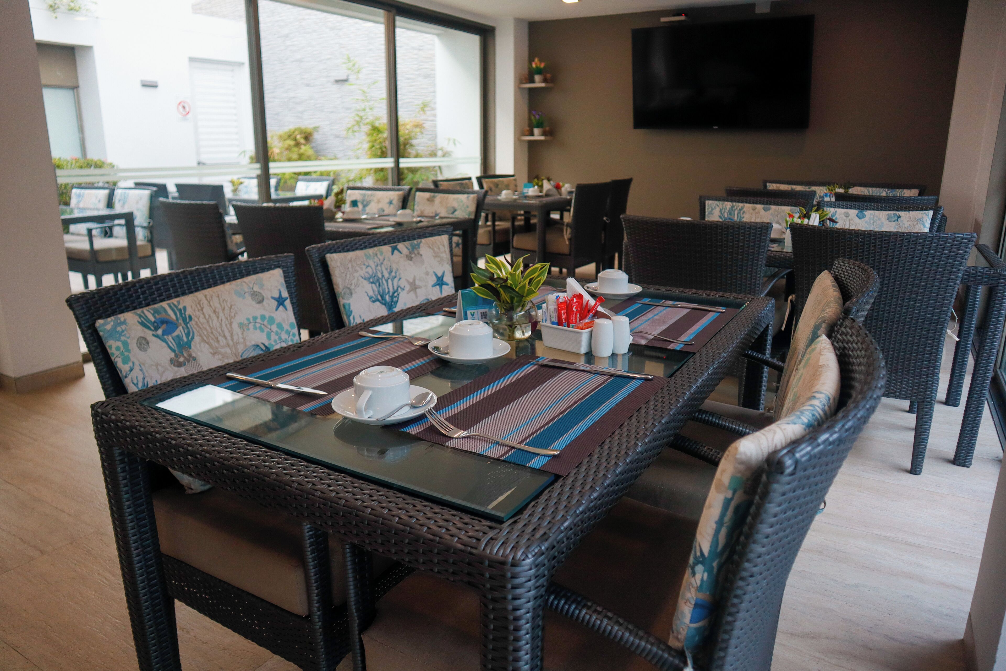 Un comedor con mobiliario de mimbre, mesas de vidrio y un televisor en la pared.