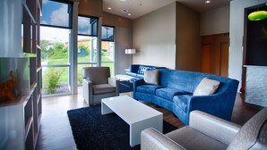Un salon moderne avec un canapé bleu, des fauteuils blancs, une table basse et une grande fenêtre donnant sur un paysage verdoyant et des bâtiments.