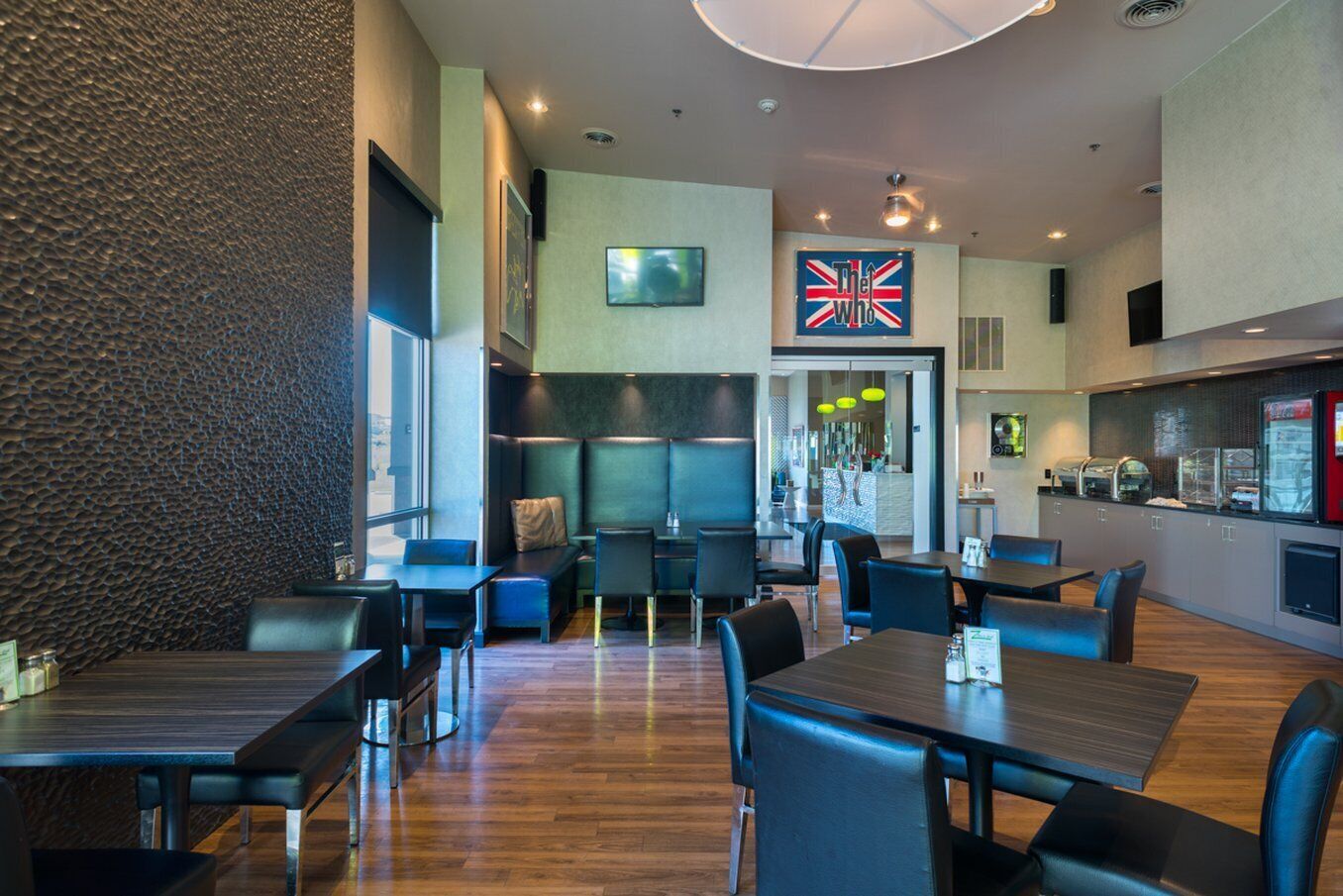 Un ristorante moderno con tavoli in legno e sedie nere, un cartello con la Union Jack e un'area cucina con un frigorifero rosso.