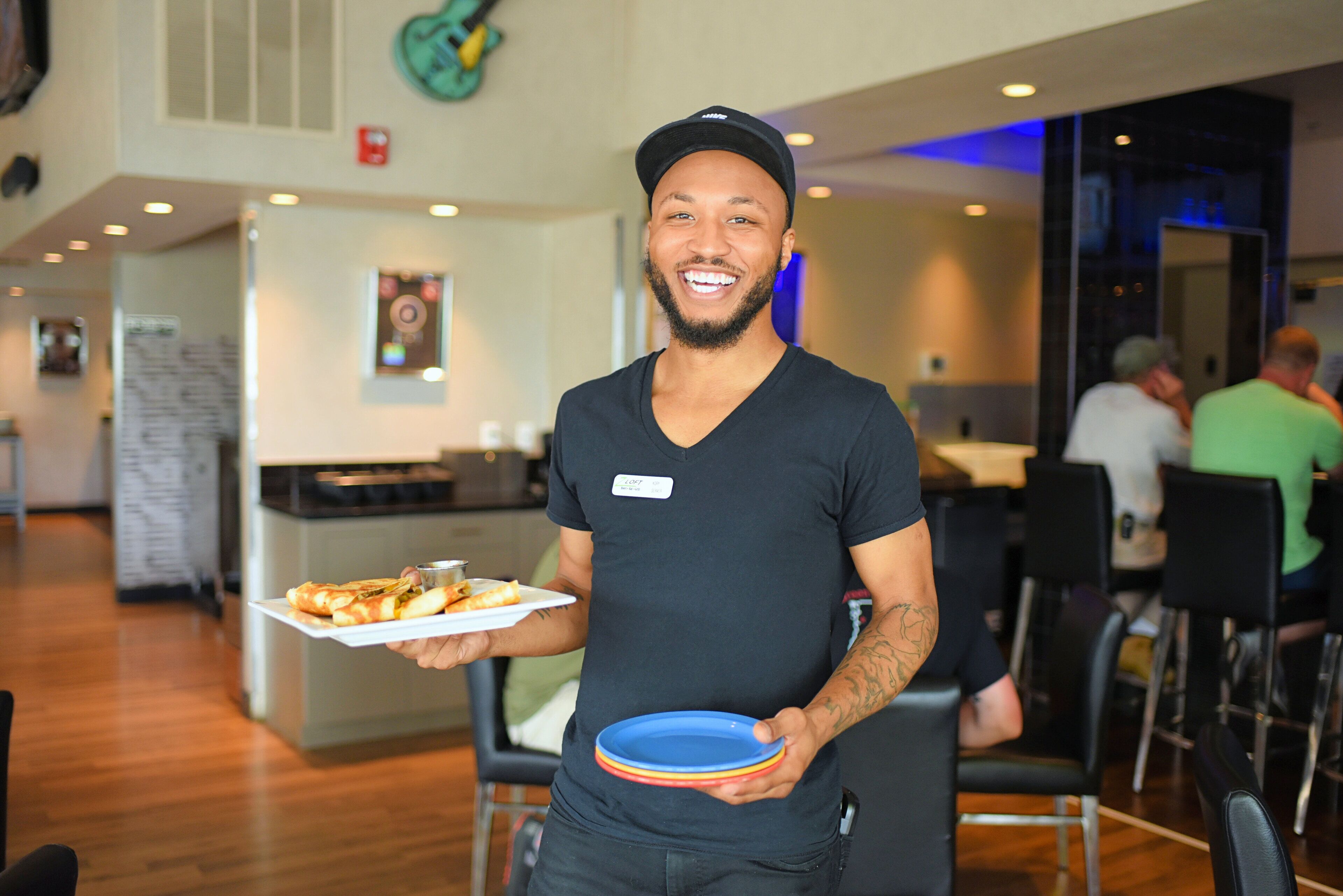 A person holding a plate of food and a stack of plates in a modern dining area with wooden flooring and contemporary furniture.