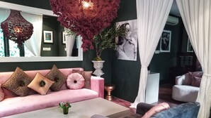 Una sala de estar moderna con un sofá rosa, un sofá gris y una mesita de centro. Hay una planta decorativa grande y un jarrón blanco sobre la mesita. La habitación tiene una pared de acento verde y cortinas blancas.