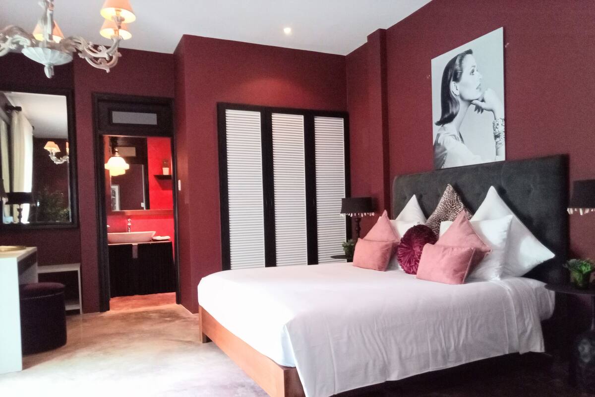 Une chambre à coucher avec un lit, une commode, un miroir, un lustre et une salle de bain éclairée en rouge.