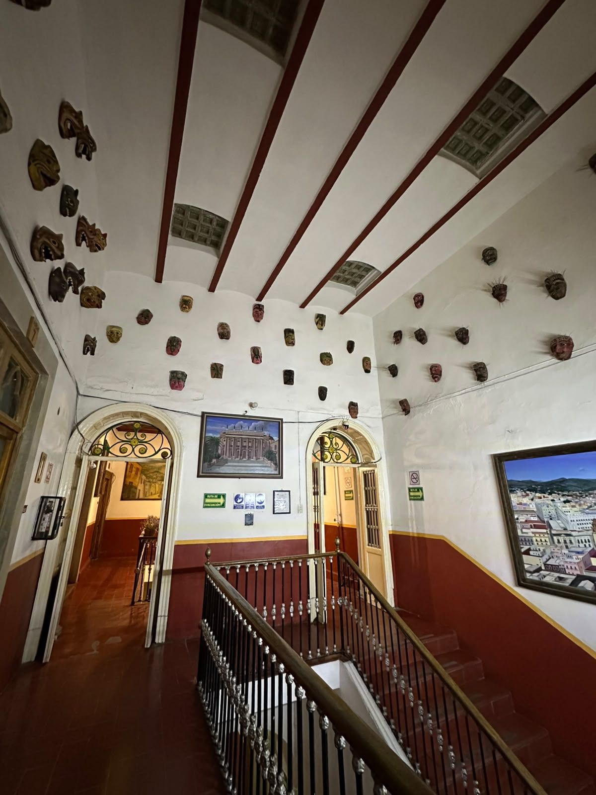 Un pasillo con una escalera, una pared decorada con numerosas máscaras y un cuadro de un edificio.