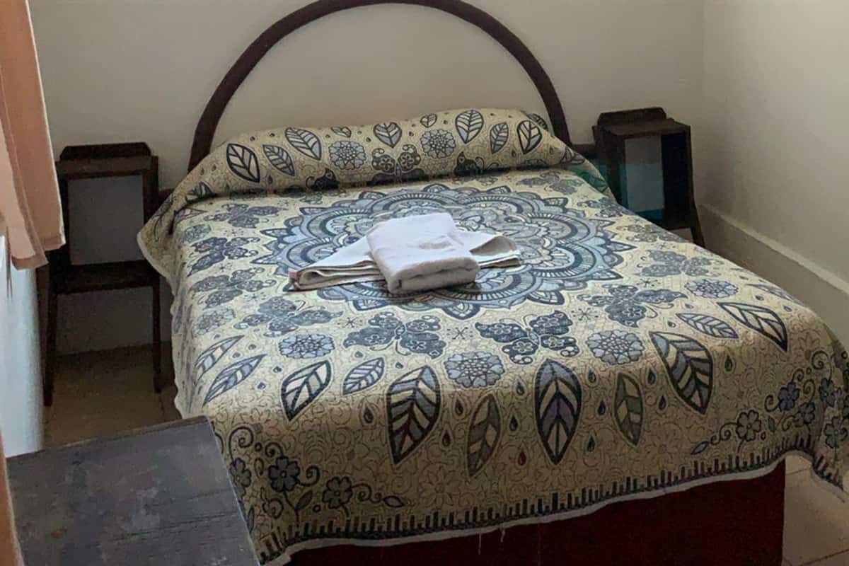 Una cama pequeña y ordenada con una colcha estampada, una mesita de noche con lámpara y una silla en una habitación acogedora.