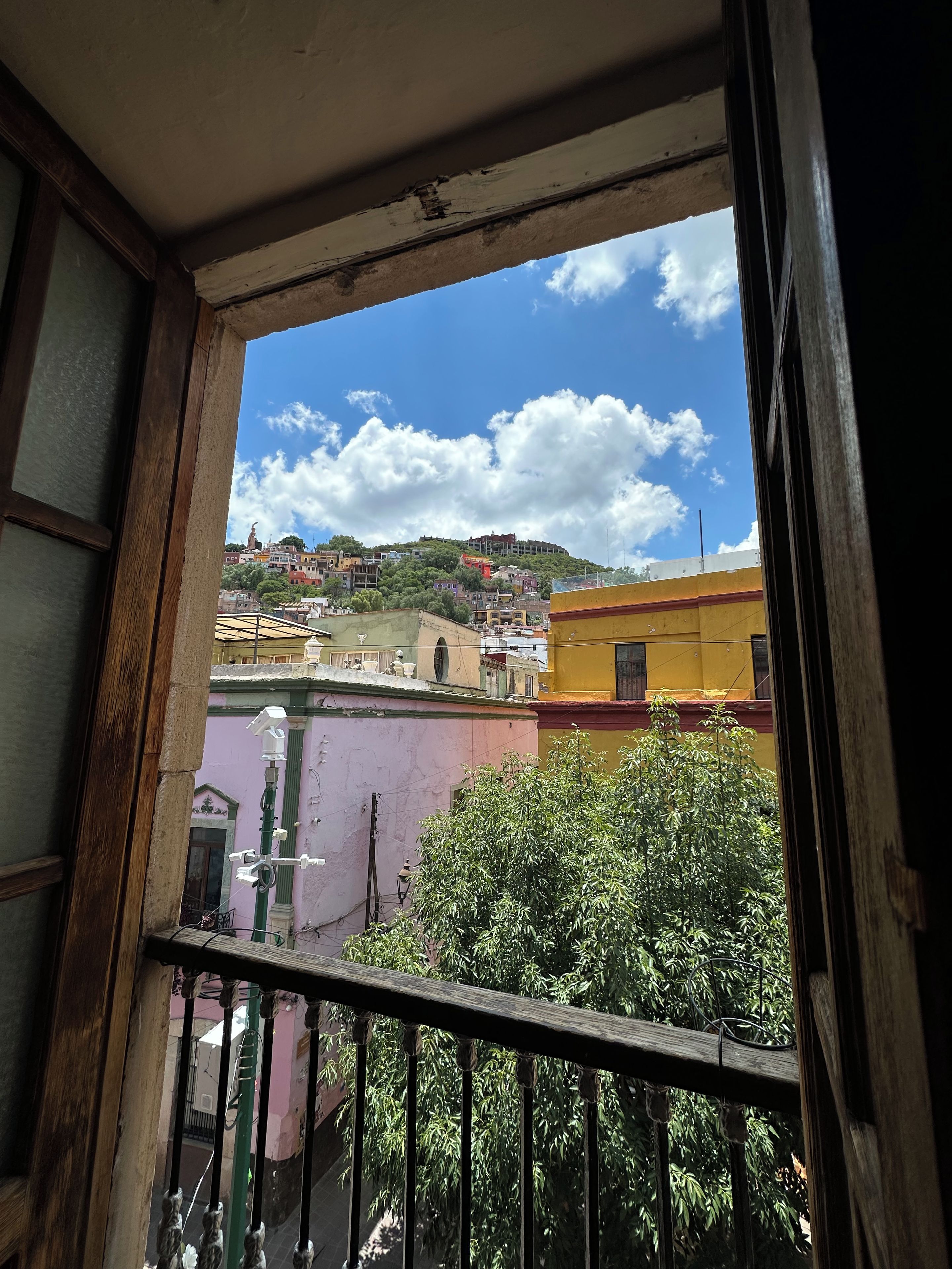 Une vue depuis une fenêtre donnant sur un balcon, avec un panorama sur la ville, ses bâtiments et une colline.