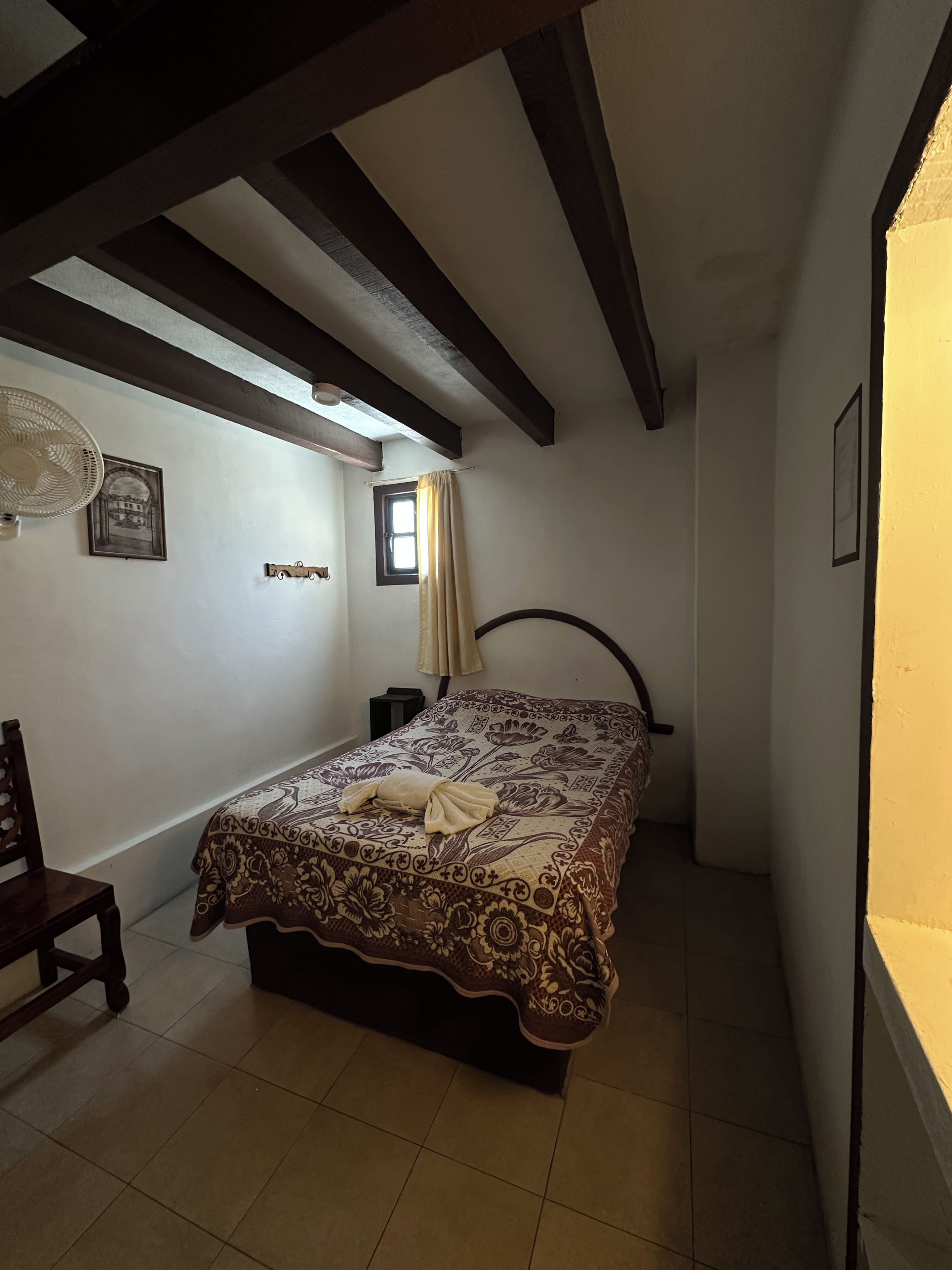 A bedroom with a bed, a chair, a small table, a fan, and a window with curtains.