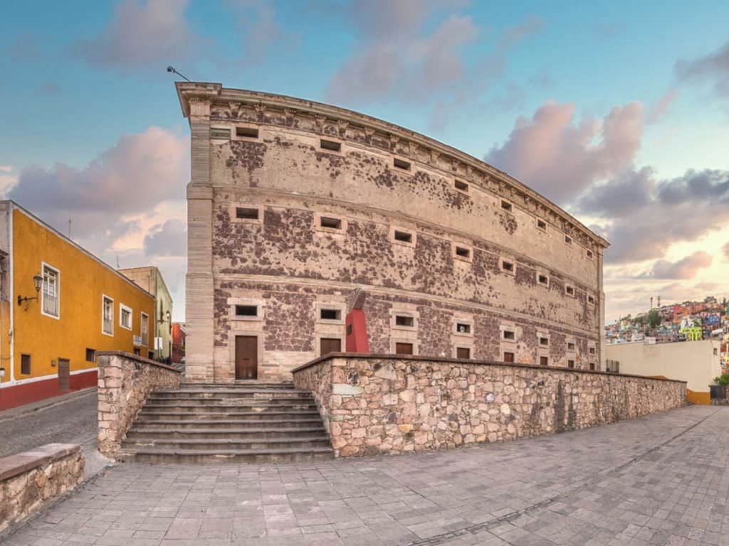 A historic building with a curved facade, a stone staircase leading to the entrance, and a paved ground.