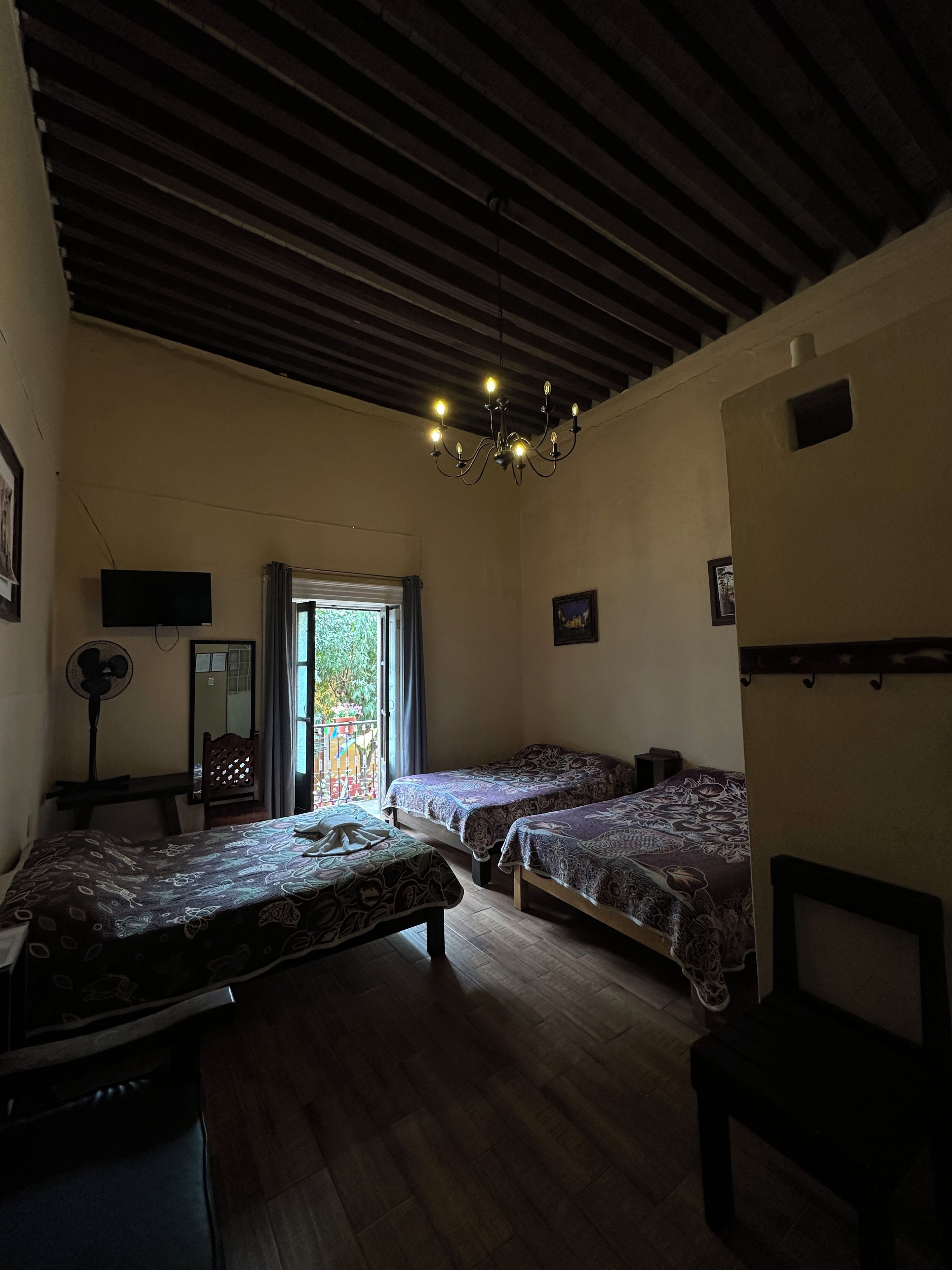 Ein Zimmer mit Holzboden, zwei Betten mit gemusterten Laken, einem Schreibtisch mit Stuhl, einem Standventilator und einem Fernseher an der Wand.