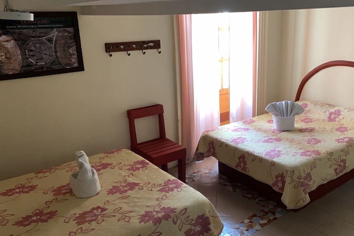 A bedroom with two beds, a chair, a small table, and a wall-mounted shelf.