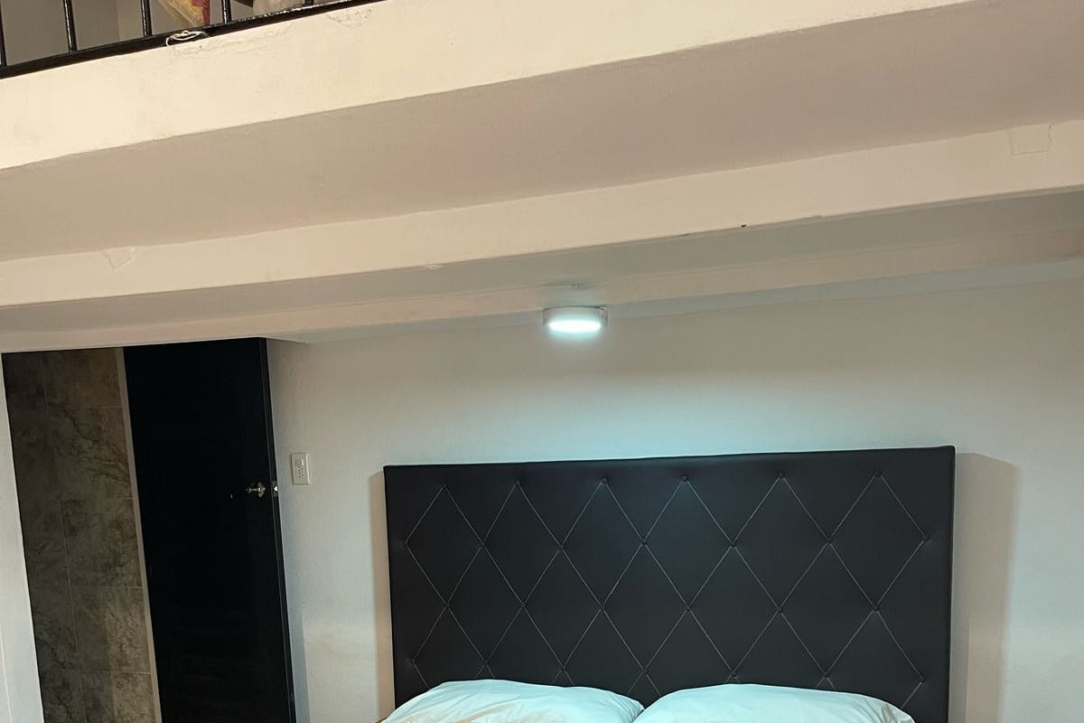 Una cama bien hecha con una colcha naranja y dos almohadas, suelo de madera, una mesita y un cabecero negro.