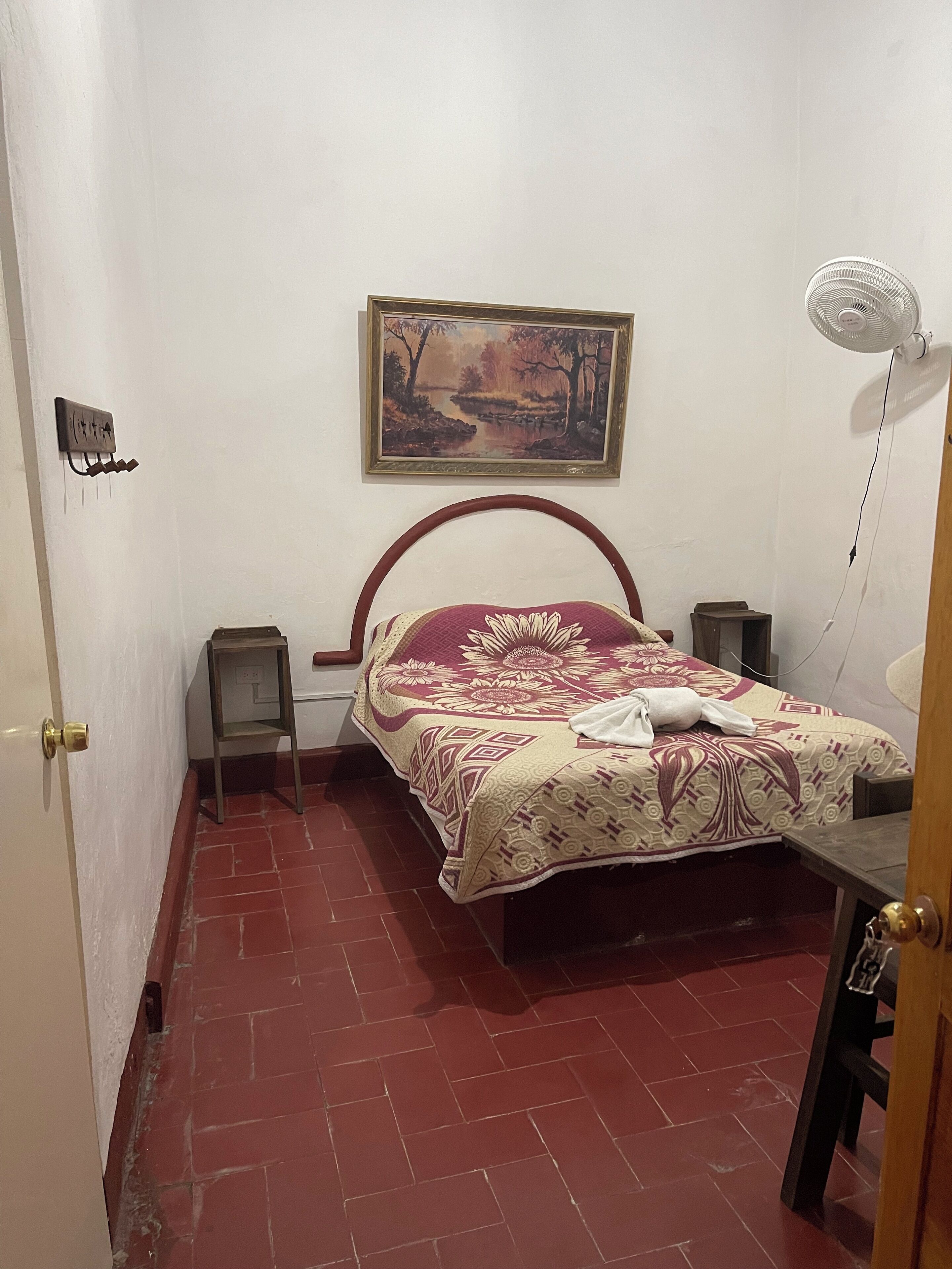 Un dormitorio con cama, un cuadro en la pared, un ventilador de pie y una mesita.