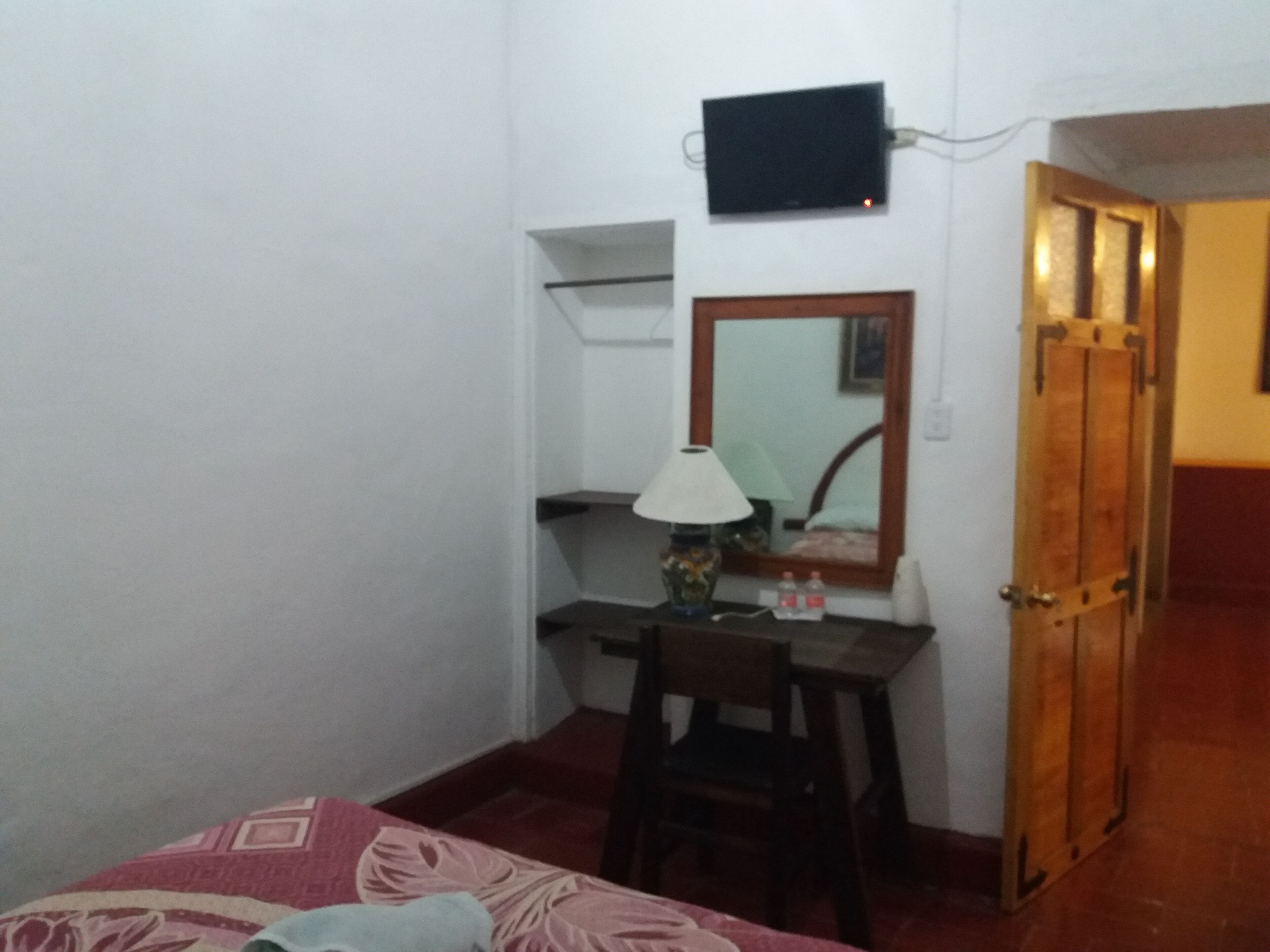 A small room with a bed, a desk with a chair, a lamp, a mirror, and a television mounted on the wall.