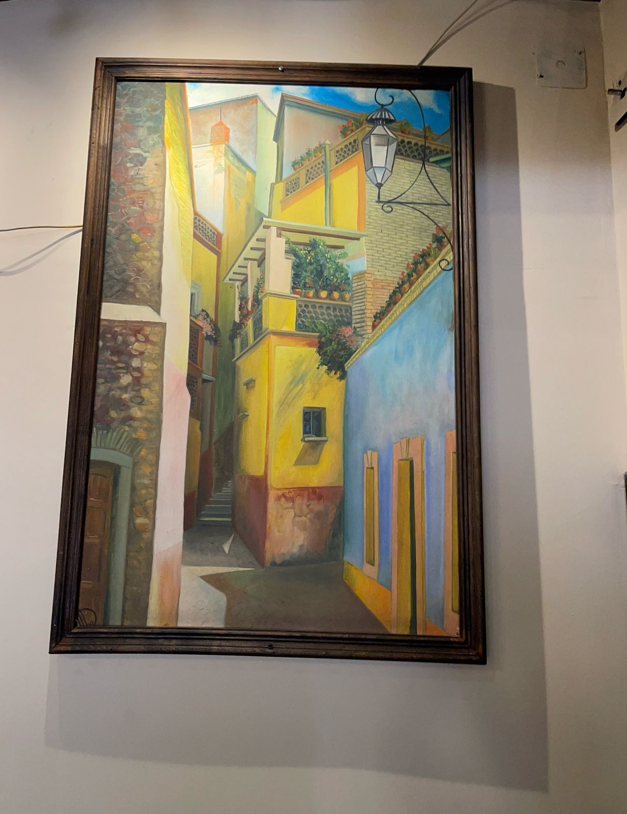 A framed painting of a narrow street with colorful buildings, a lamp post, and a staircase.