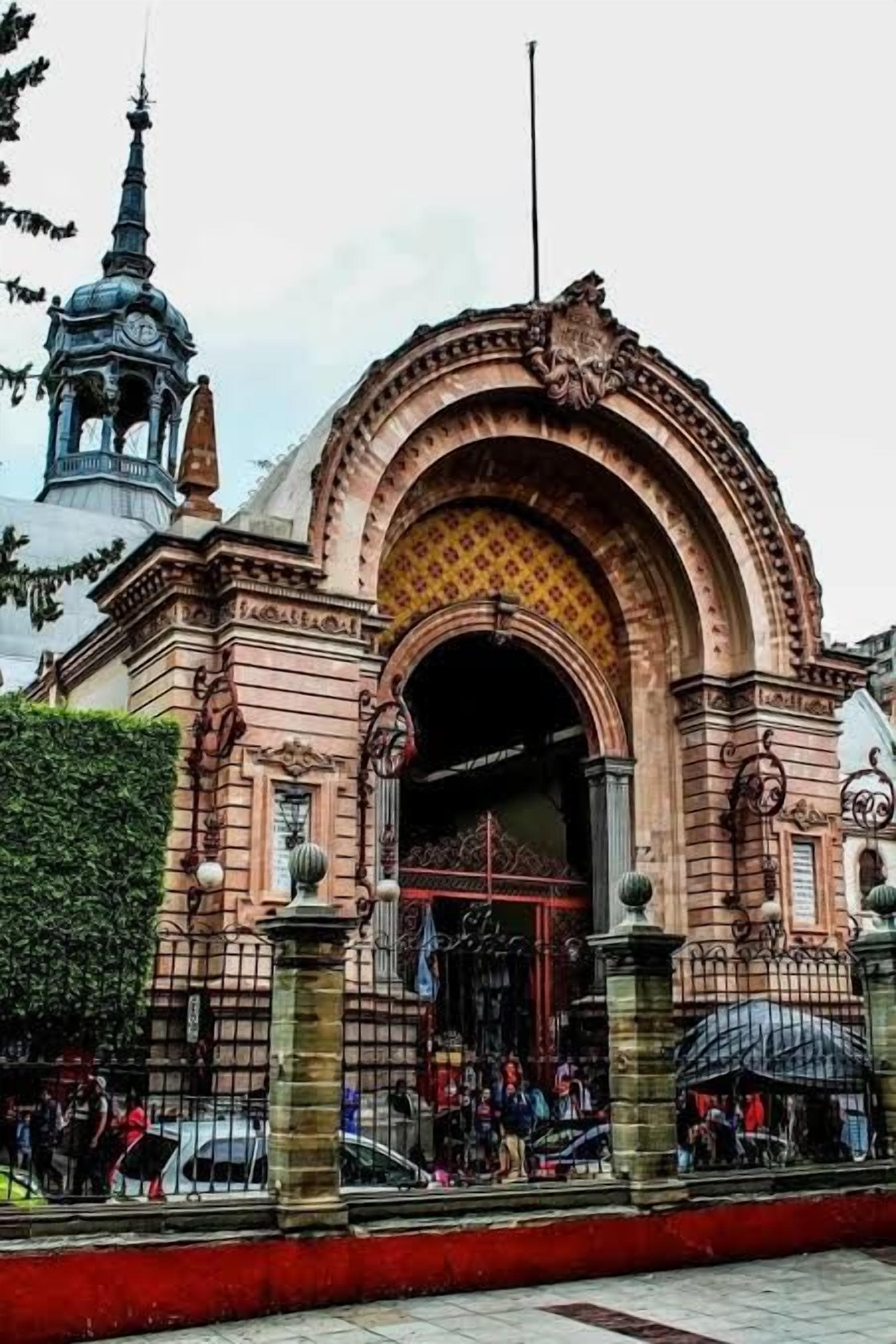Un edificio histórico con una entrada ornamentada, una torre del reloj y un área vallada con gente.