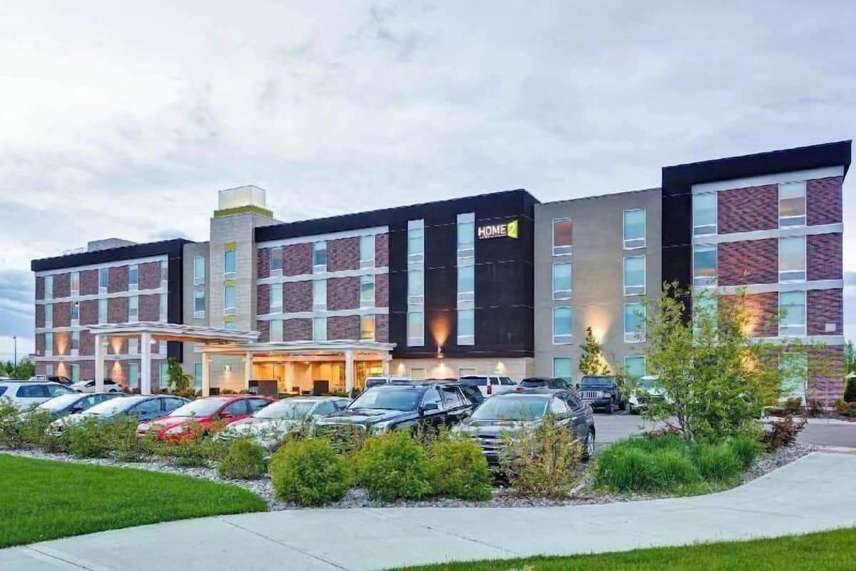 A multi-story hotel with a modern facade, a parking lot with several cars, and a well-maintained lawn area.
