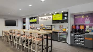 A modern coffee shop with a long counter, multiple chairs, a TV, and a coffee and tea station.