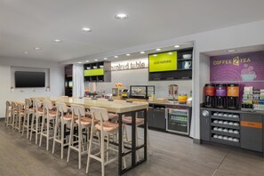 Una cafetería moderna con una barra alargada, varias sillas, un televisor y una estación de café y té.