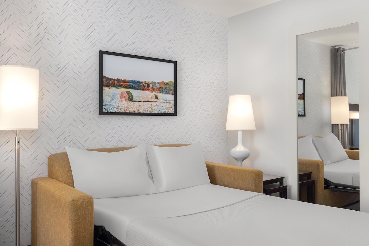 A hotel room with a bed, two bedside lamps, a large mirror, and a framed picture on the wall.
