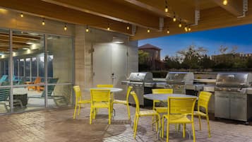 An outdoor patio with yellow chairs and tables, a grill station, and a glass wall overlooking a building.