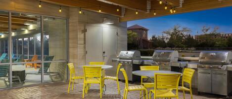 An outdoor patio with yellow chairs and tables, a grill station, and a glass wall overlooking a building.