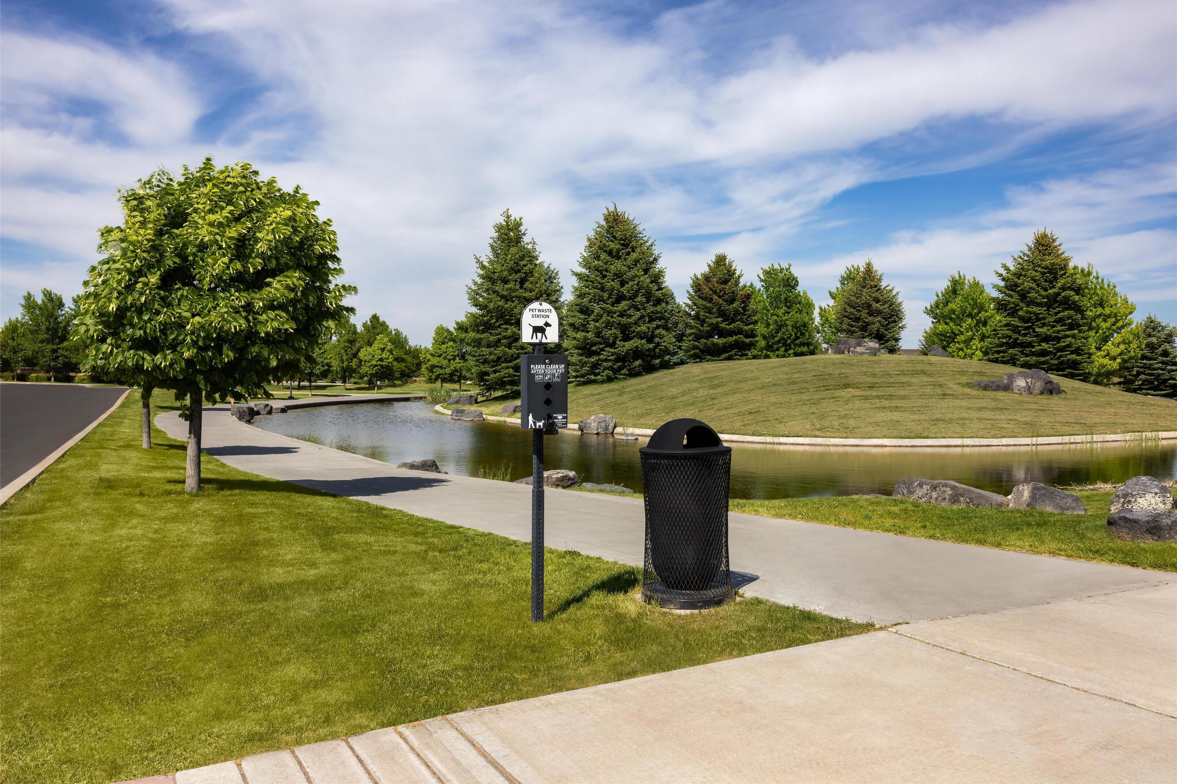 A pet waste station with a sign, a trash can, a pond, and a grassy area with trees.