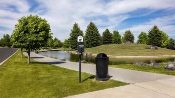 A pet waste station with a sign, a trash can, a pond, and a grassy area with trees.