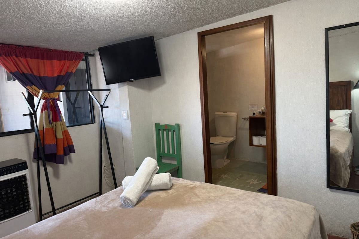 Un dormitorio con cama, televisor montado en la pared, una silla verde y un baño visible a través de una puerta abierta.