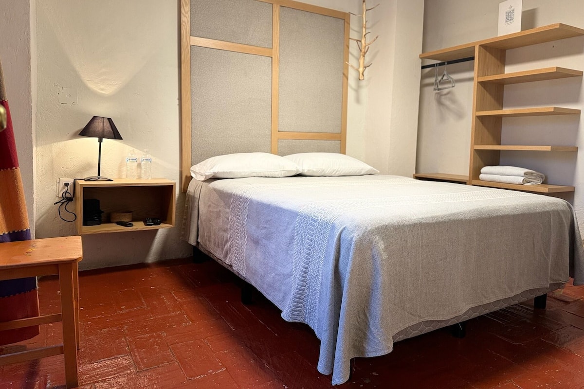 Un dormitorio ordenado con cama, mesitas de noche, una lámpara y un estante con toallas.