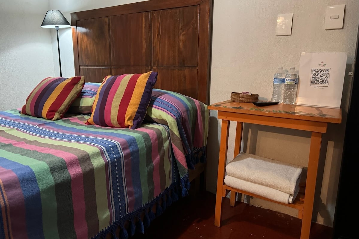 Una habitación con cama, cabecero de madera, mesita de noche con una toalla, lámpara y un código QR en la pared.