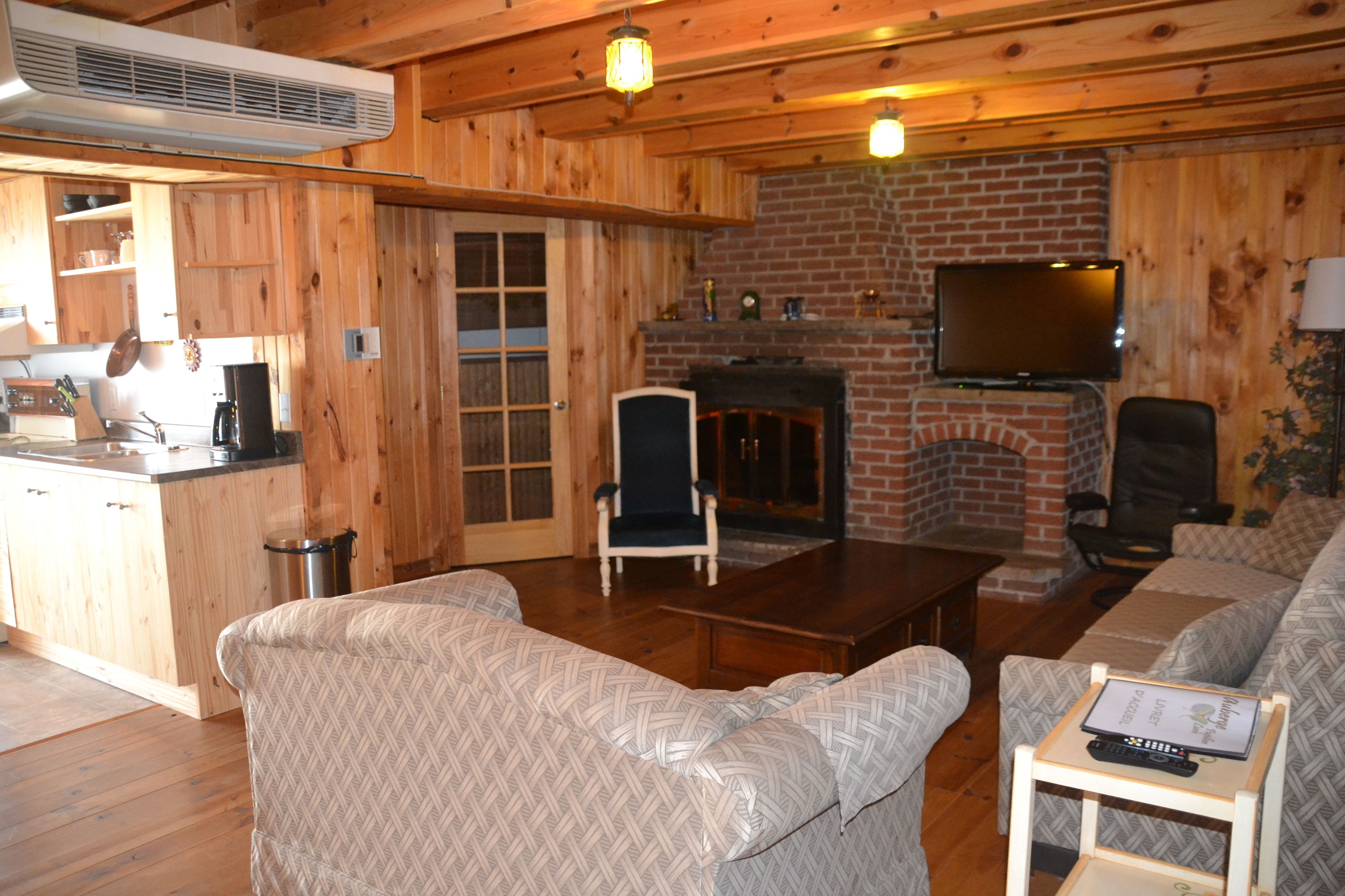 A cozy cabin interior with wooden walls, a brick fireplace, a television mounted above it, and a dining area with a table and chairs.