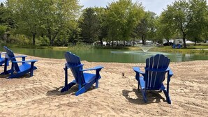 Una playa arenosa con tres sillas Adirondack azules mirando un lago tranquilo, rodeada de árboles y vehículos recreativos al fondo.