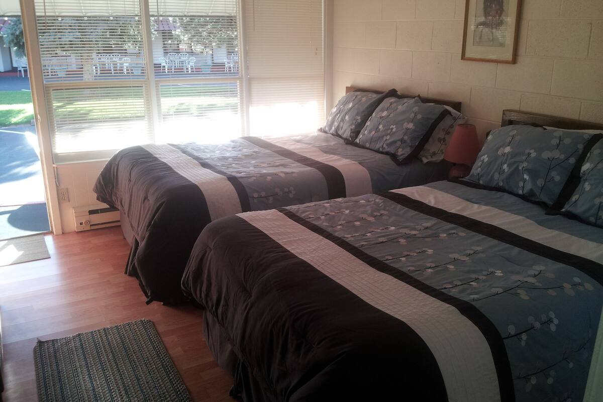 A bedroom with two beds, a window with blinds, a framed picture on the wall, and a wooden floor.