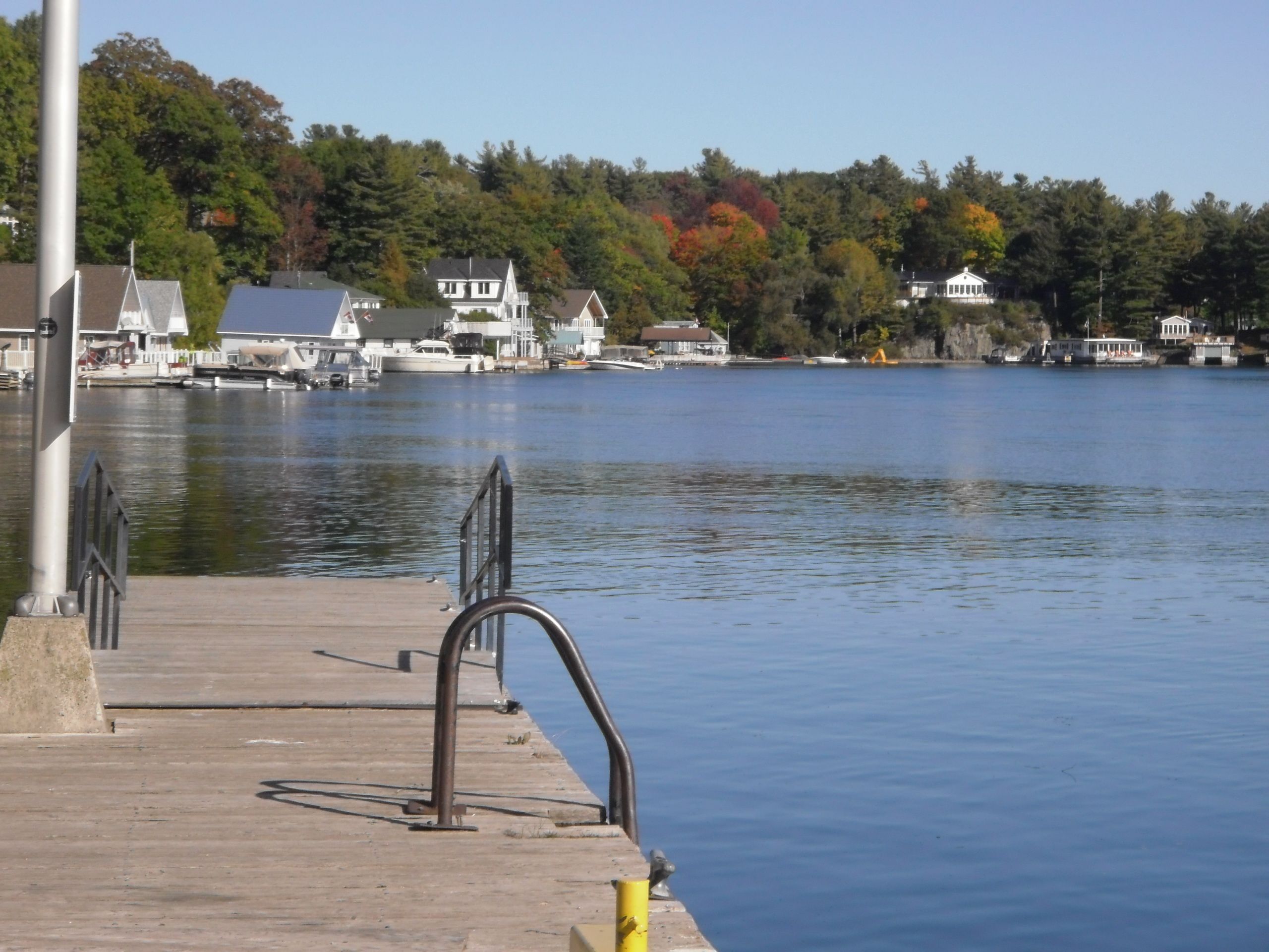 A waterfront dock with a metal railing, a concrete walkway, and houses in the background.