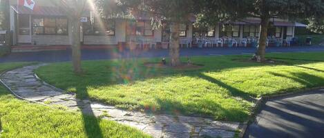 A grassy area with a stone pathway leading to a building with a Canadian flag. There are outdoor tables and chairs under a covered area.