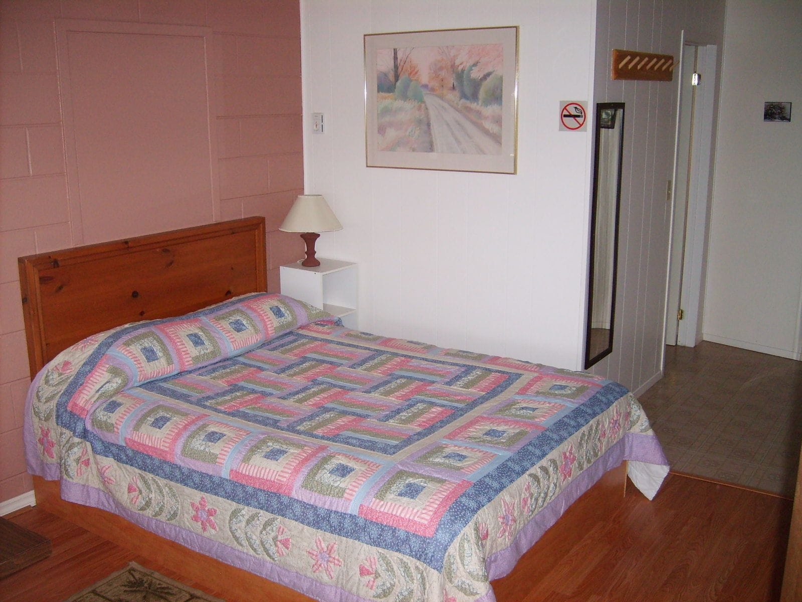 Un dormitorio con una cama de madera, una mesita de noche con lámpara, un cuadro enmarcado en la pared y una puerta con letrero de 'no fumar'.