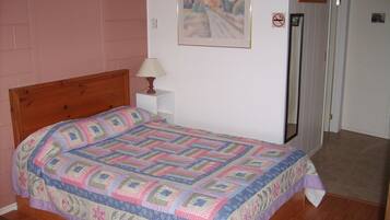Un dormitorio con una cama de madera, una mesita de noche con lámpara, un cuadro enmarcado en la pared y una puerta con letrero de 'no fumar'.