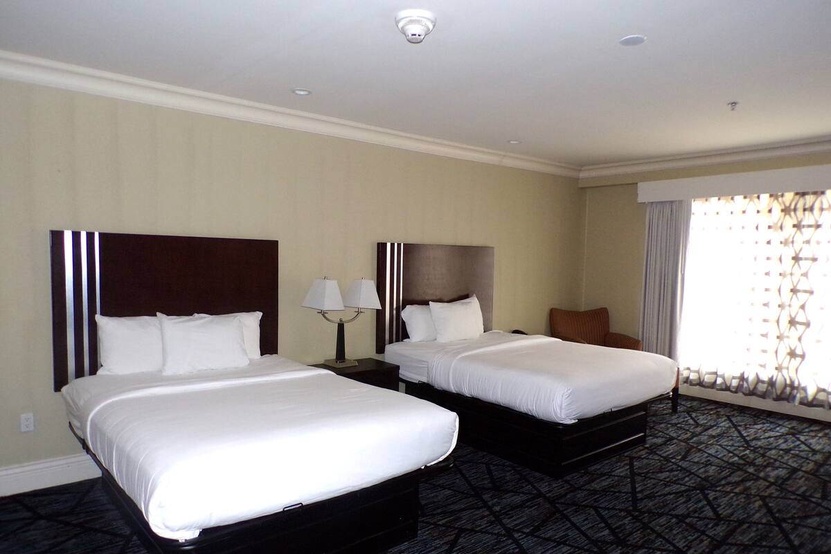 A hotel room with two beds, a nightstand with lamps, a chair, and a large window with curtains.