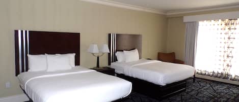 Habitación de hotel con dos camas, una mesita de noche con lámparas, una silla y un amplio ventanal con cortinas.