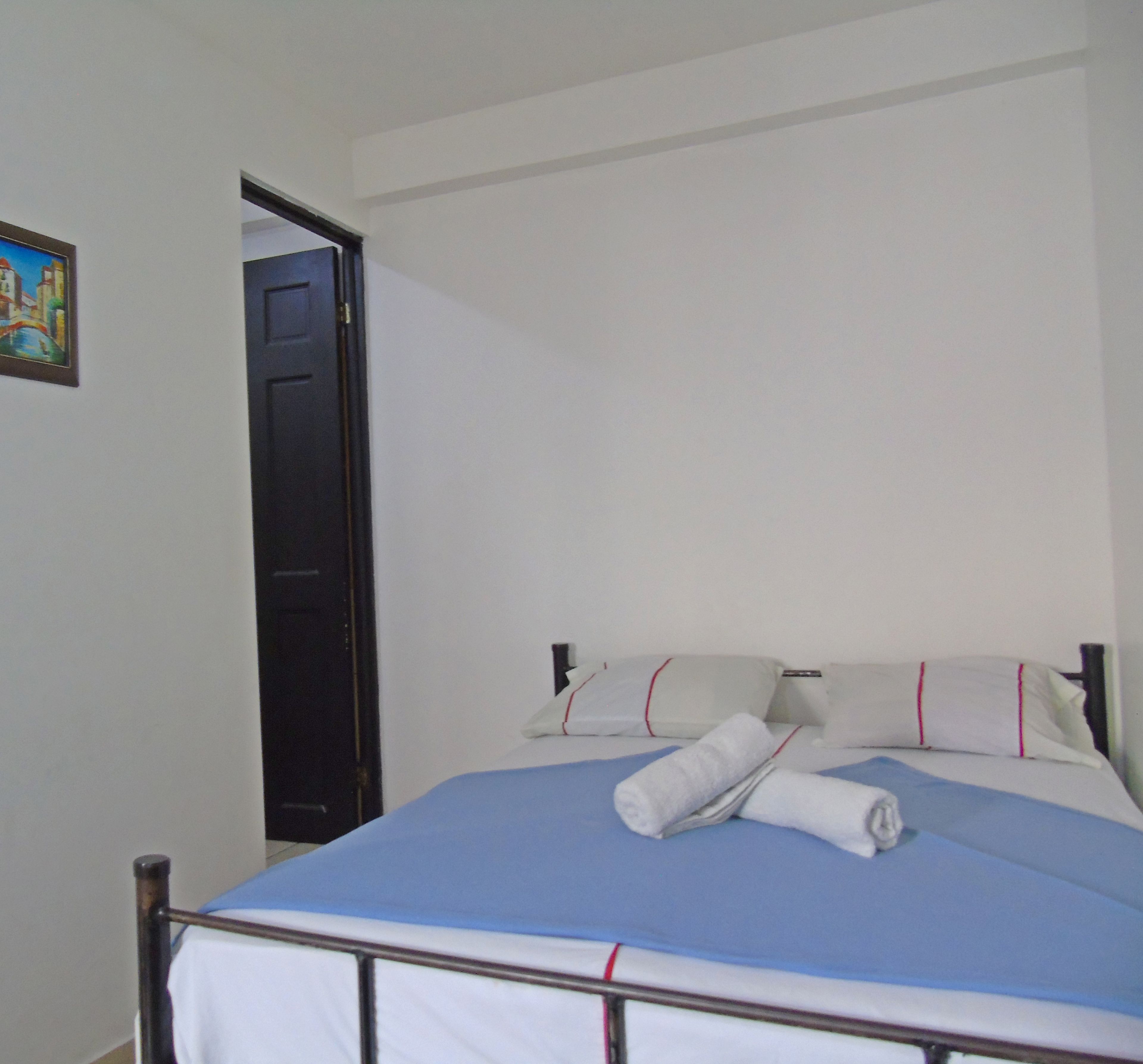 A single bed with a blue mattress and white bedding, a white pillow with red trim, and a rolled white towel on the bed.