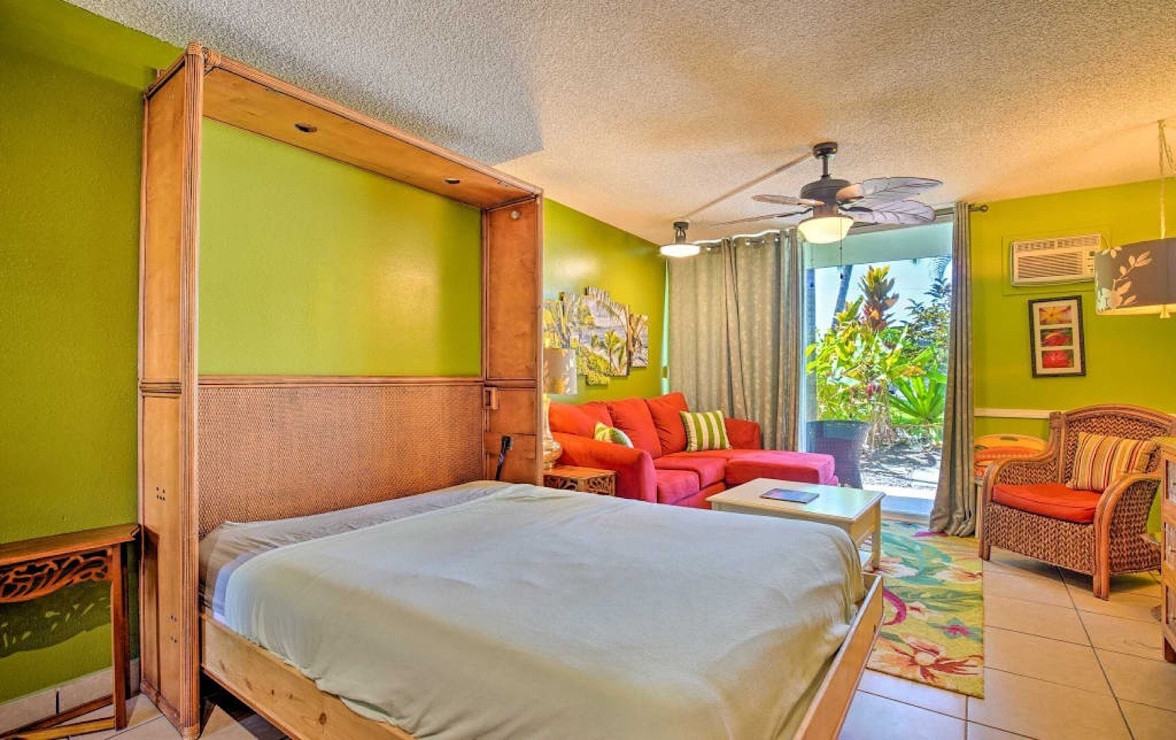 A room with a bed, a red sofa, a wicker chair, and a ceiling fan.