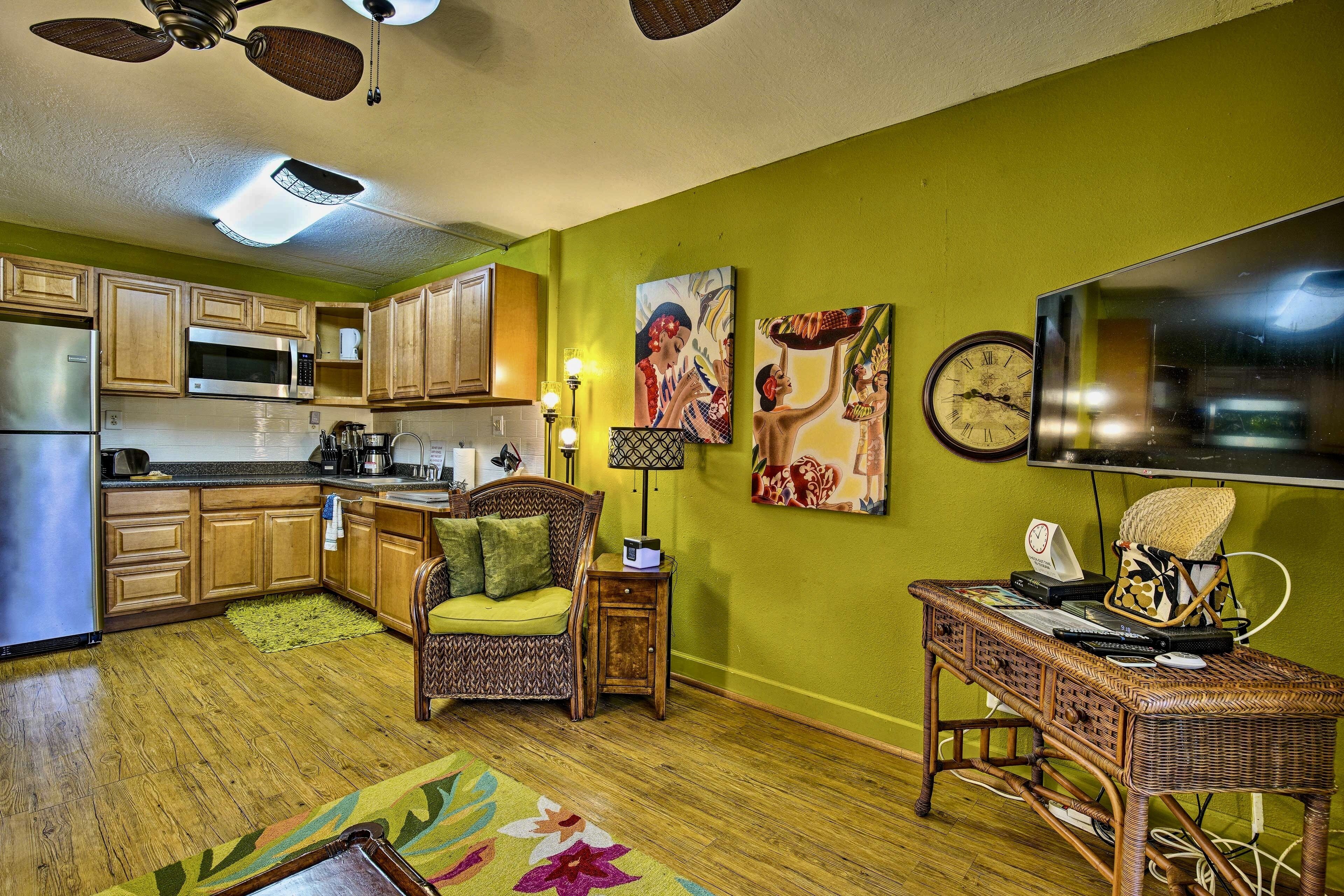 Una cocina con armarios de madera, un microondas y un refrigerador. Hay una zona de comedor con una silla y una mesa. Las paredes están pintadas de verde, y hay un televisor de pantalla plana grande en la pared.