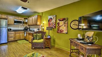 Una cocina con armarios de madera, un microondas y un refrigerador. Hay una zona de comedor con una silla y una mesa. Las paredes están pintadas de verde, y hay un televisor de pantalla plana grande en la pared.