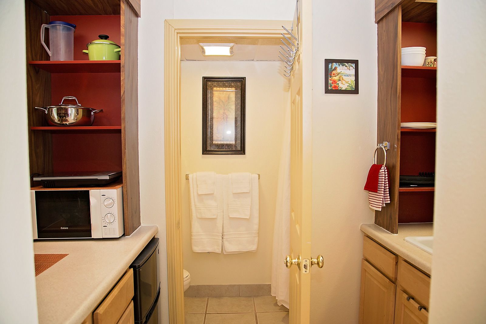 A narrow kitchen with a microwave, stove, and shelves with kitchenware. A door with a towel hanging on it is visible.