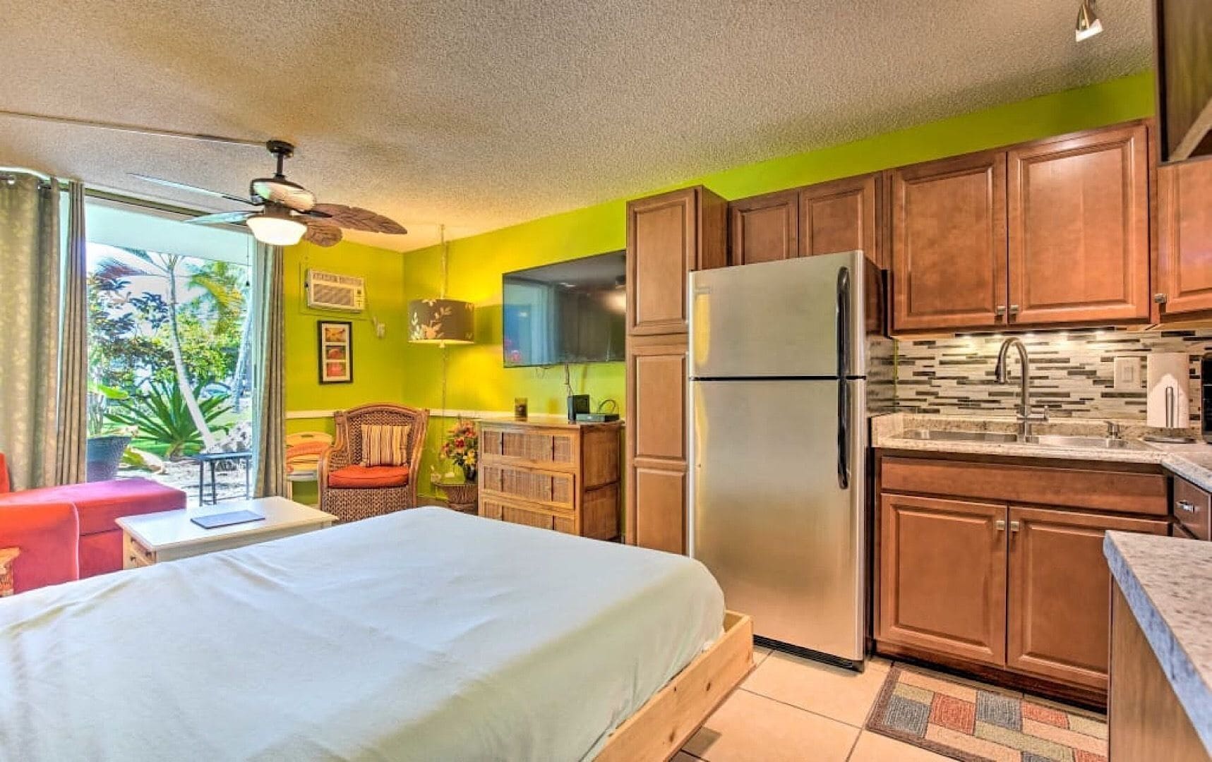 Habitación compacta con cama, un pequeño espacio para sentarse, kitchenette con refrigerador y ventana con vista a zonas verdes.