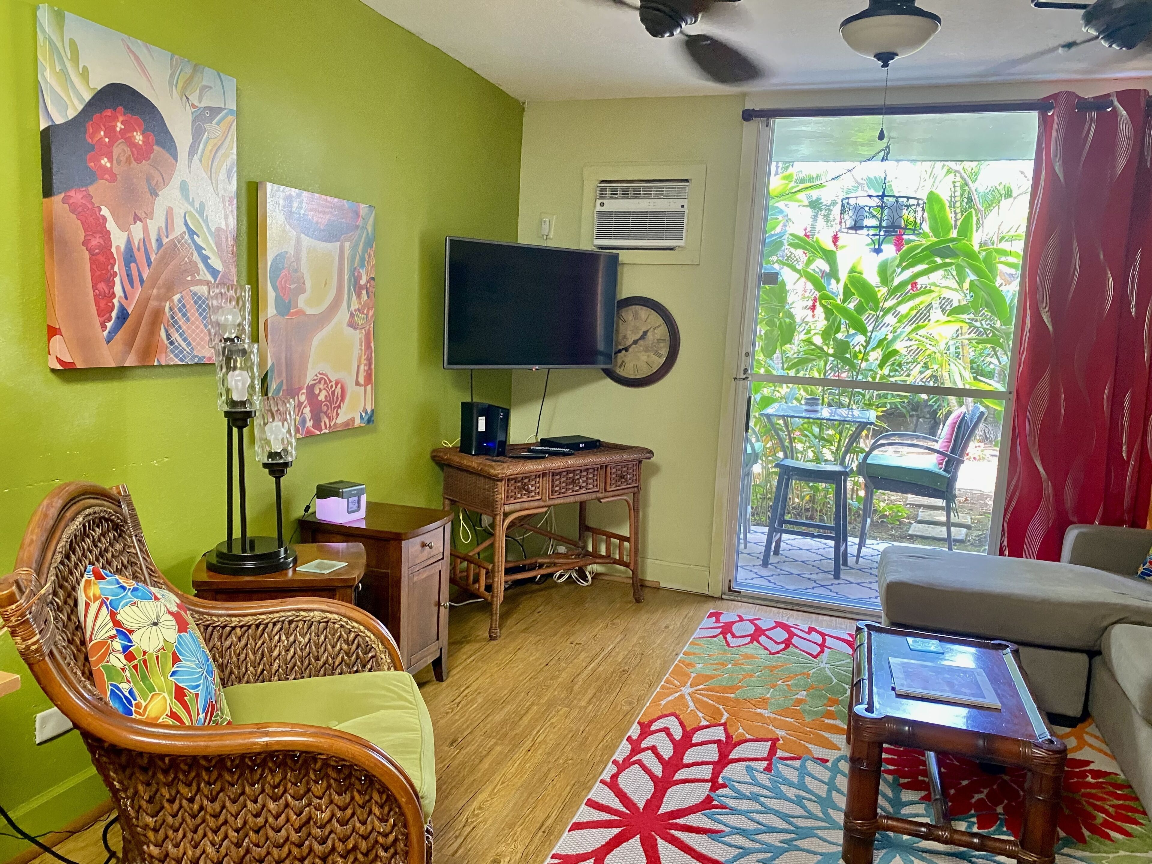 A living room with a green wall, a flat-screen TV, a wicker chair, a sofa, a small table, and a colorful rug.