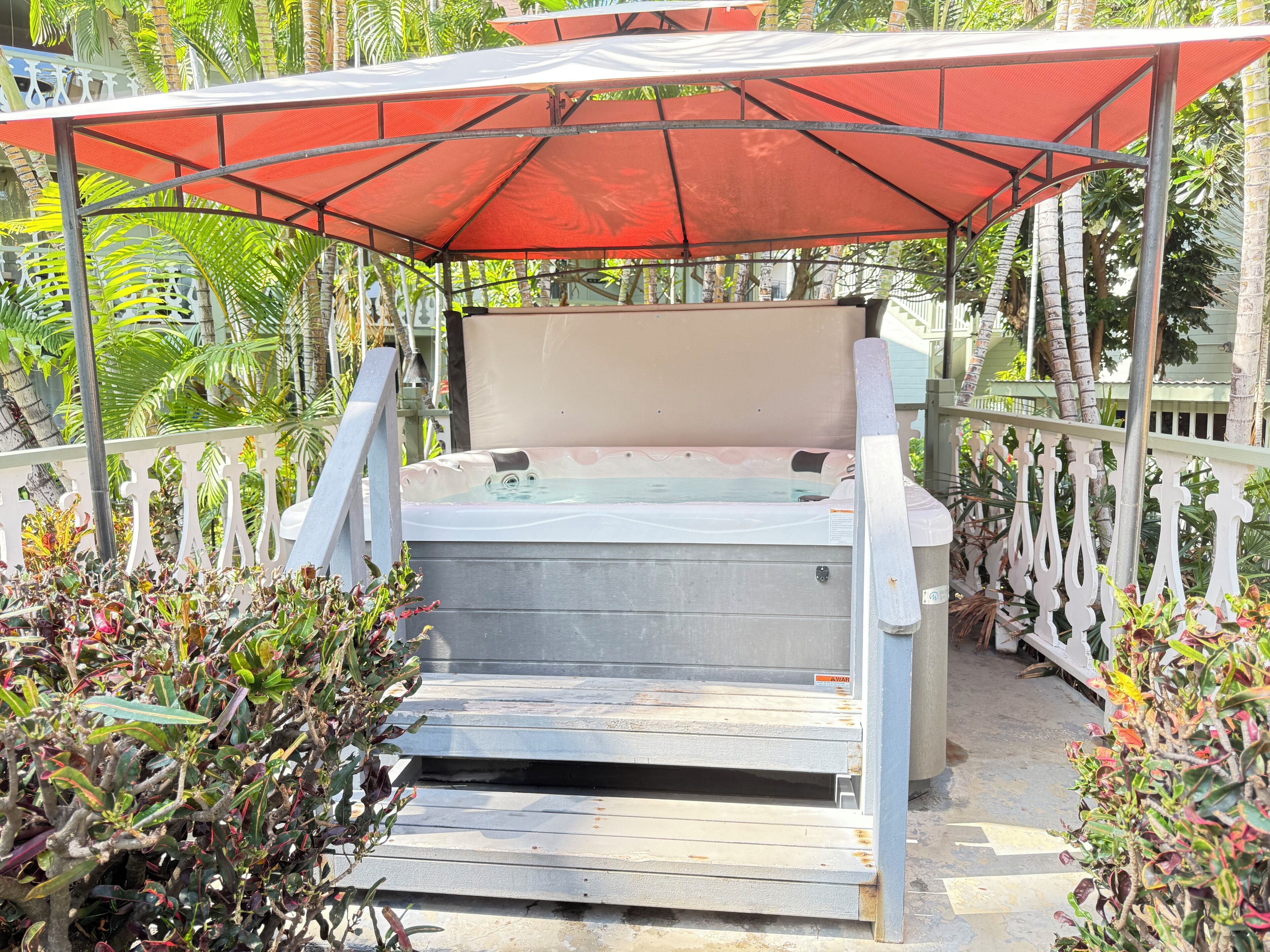 A hot tub under a red canopy, surrounded by a white fence and tropical plants.