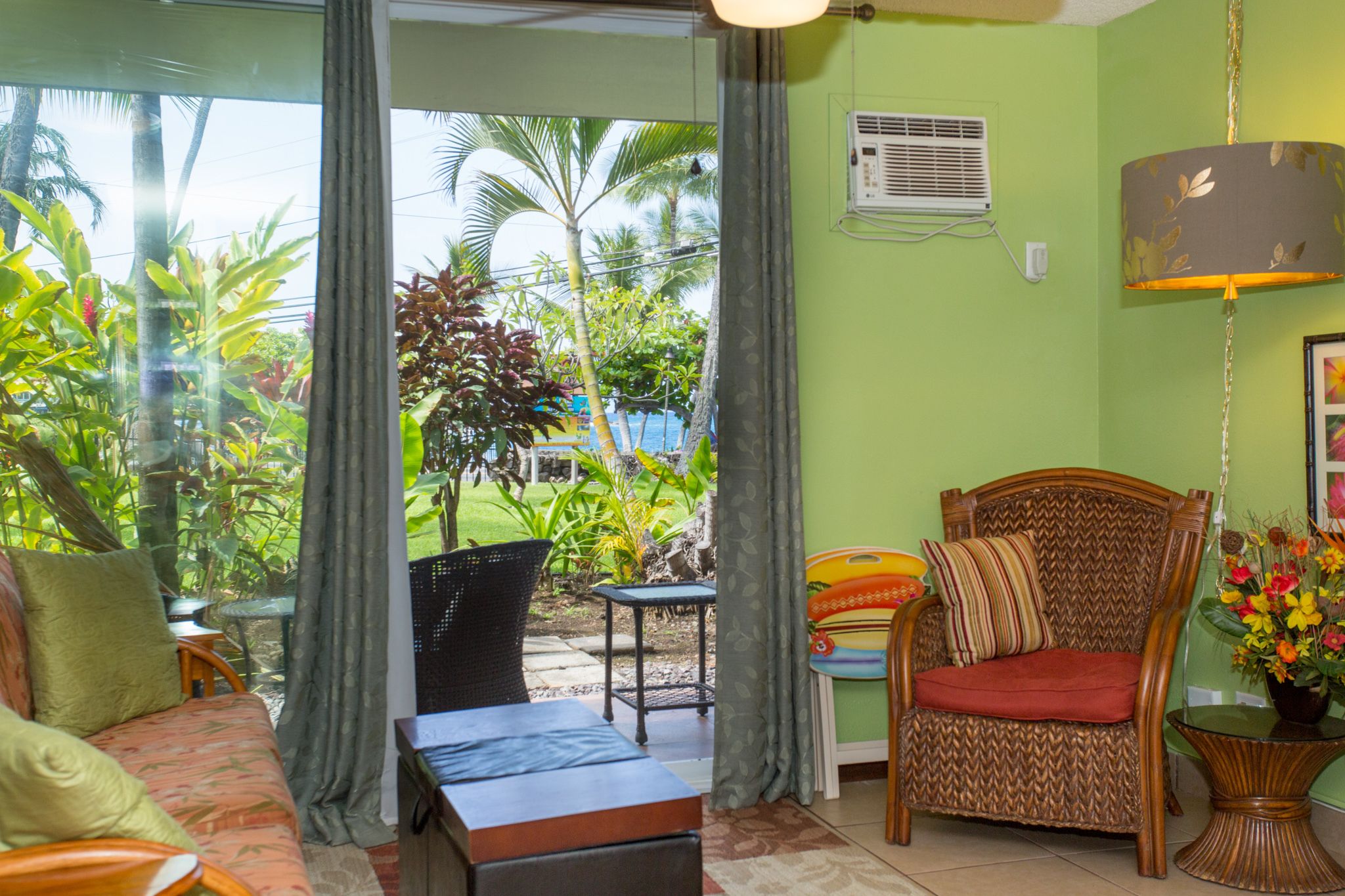 A room with a view of tropical plants and a pool, a wicker chair, a sofa, and a table.