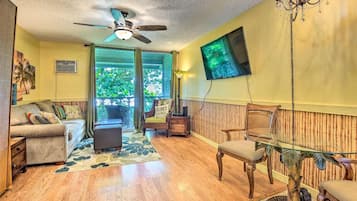 A living room with a ceiling fan, a TV mounted on the wall, a glass table, and a sofa.